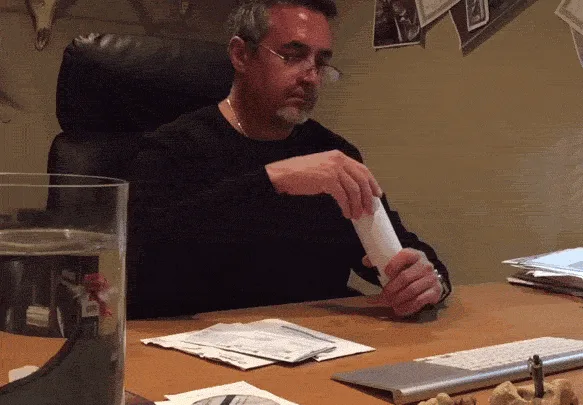 A man in glasses and a black shirt sits at a desk with a large glass container of water and papers. He holds a white…
