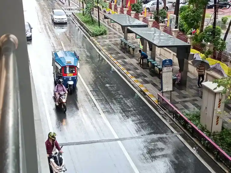 A high-angle view of a wet city street during rain, showing a blue tuk-tuk, a scooter, and a motorcycle driving past a bus…