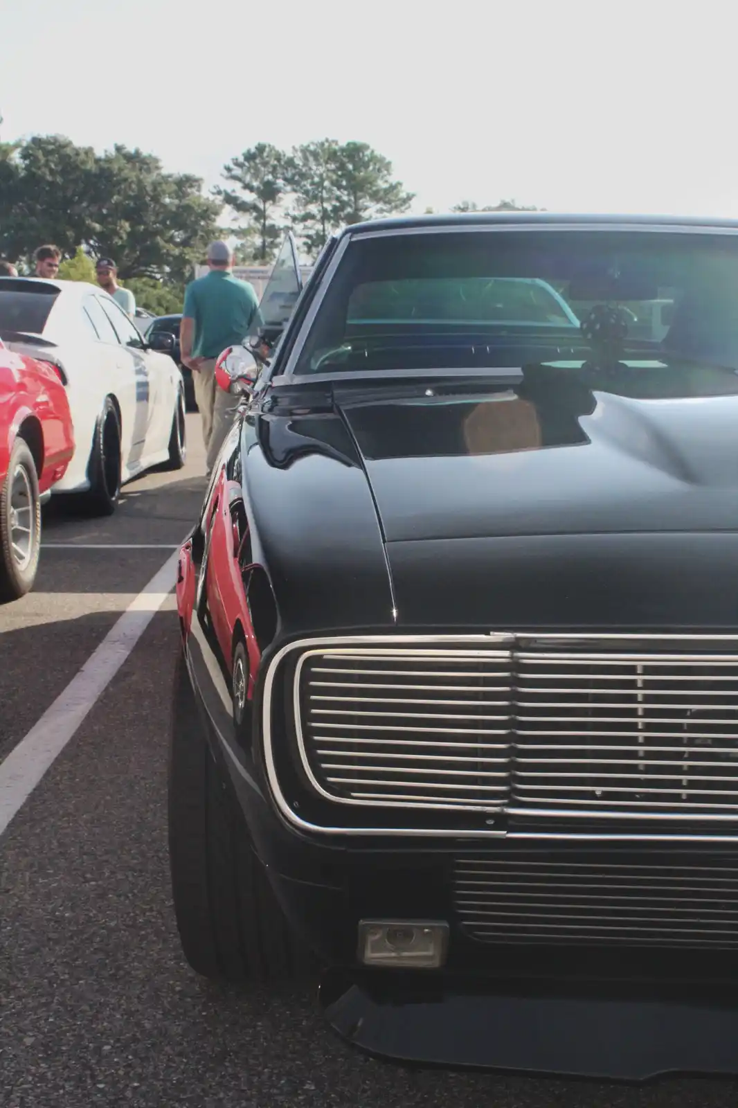 A close-up shot of a black classic muscle car with a chrome grille and hood scoop, parked at an outdoor car show with other…