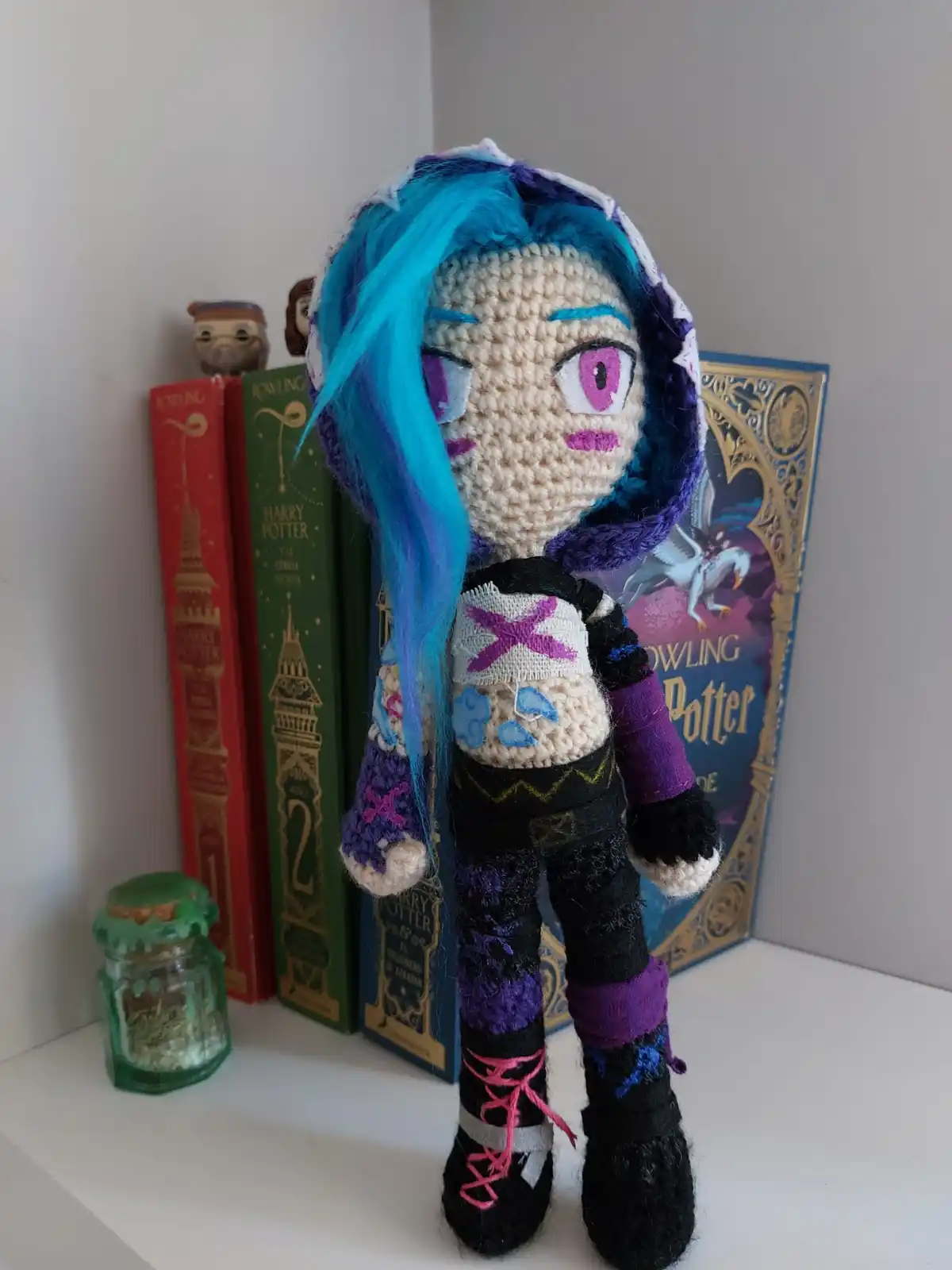 A handmade crocheted doll with blue hair and purple eyes stands on a shelf in front of Harry Potter books. The doll wears a…