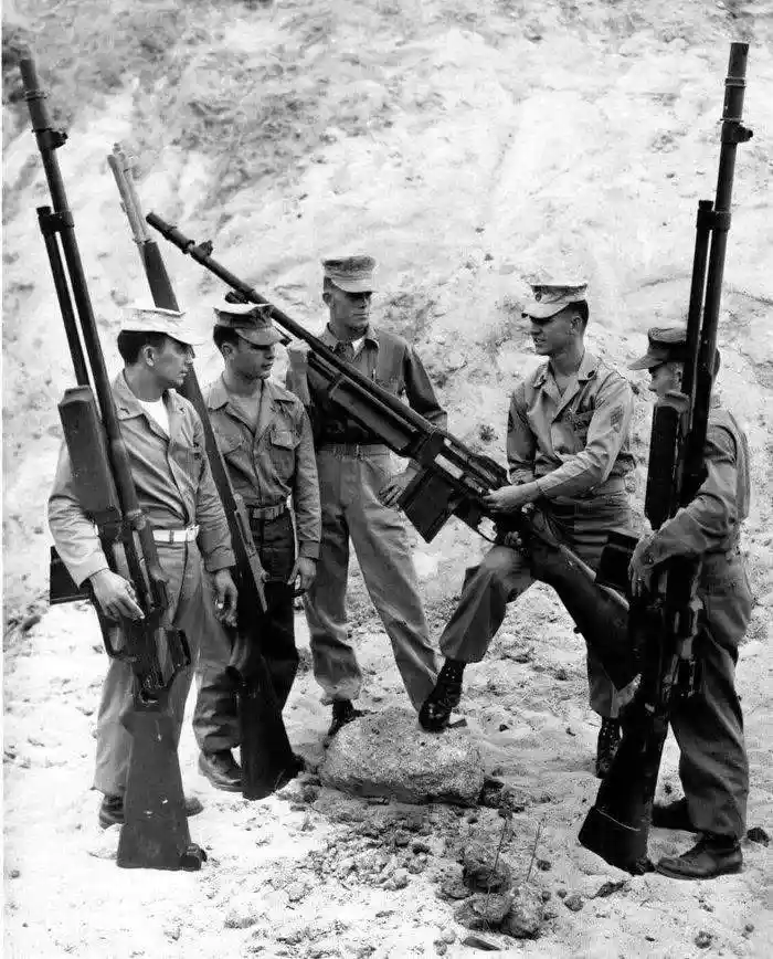 A black-and-white historical photo shows five soldiers in military uniforms standing around a large machine gun mounted on a…
