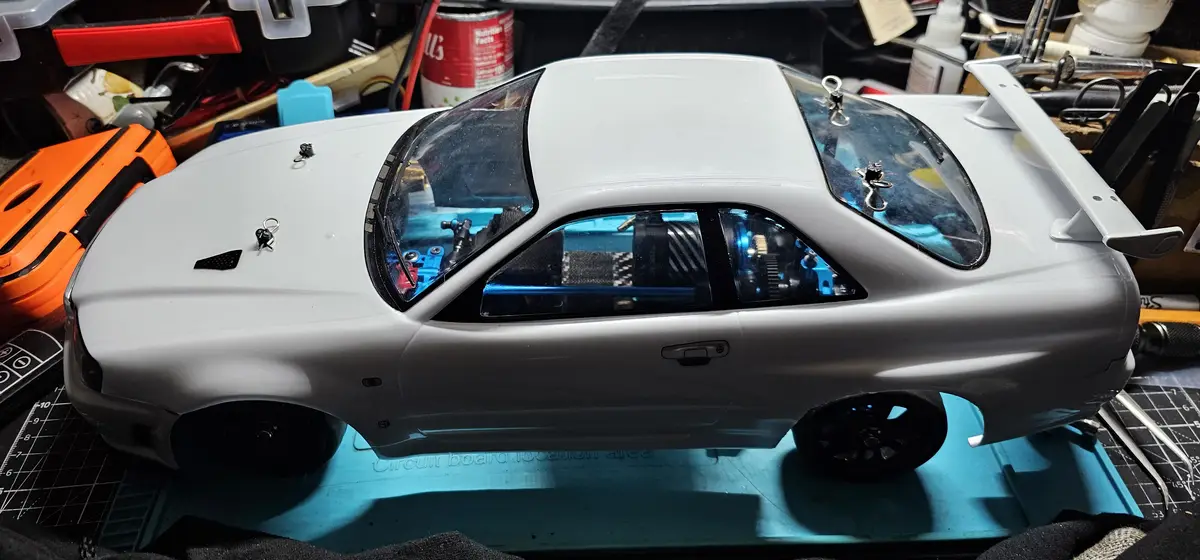 A white scale model of a sports car, likely a Nissan Skyline R32, sits on a blue work mat surrounded by tools and clutter…