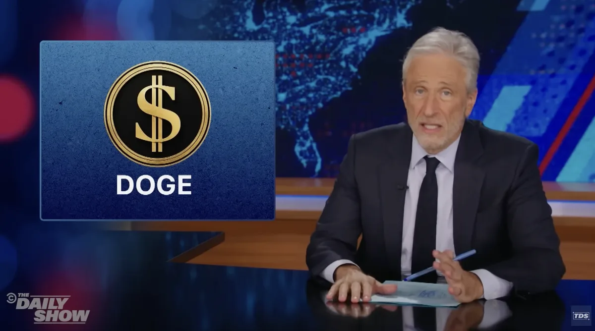 A man in a suit sits at a news desk with a graphic overlay showing a gold dollar sign inside a coin and the word 'DOGE'…