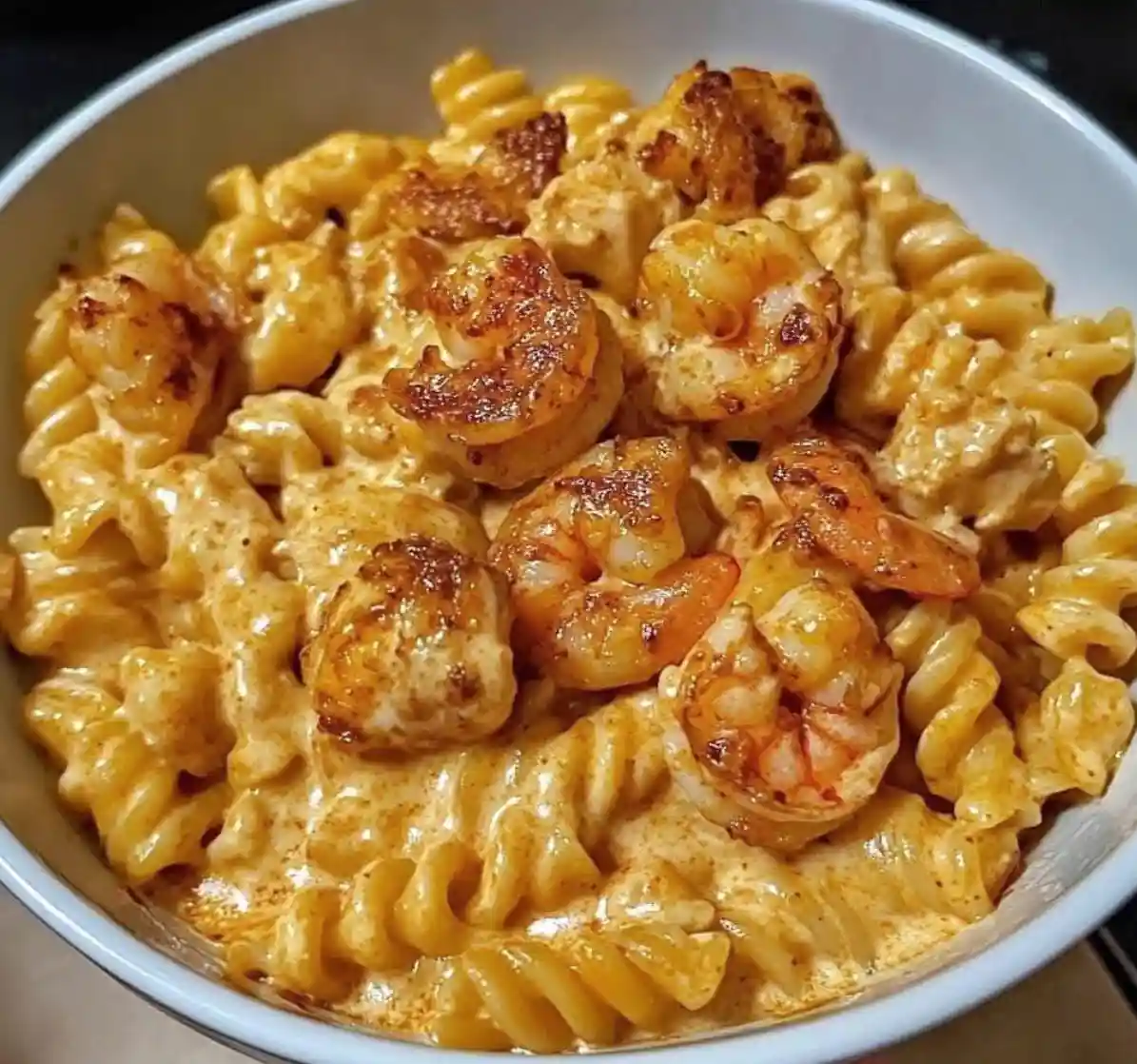 A close-up photo of a white bowl filled with creamy, orange-hued macaroni and cheese topped with seared shrimp. The pasta is…
