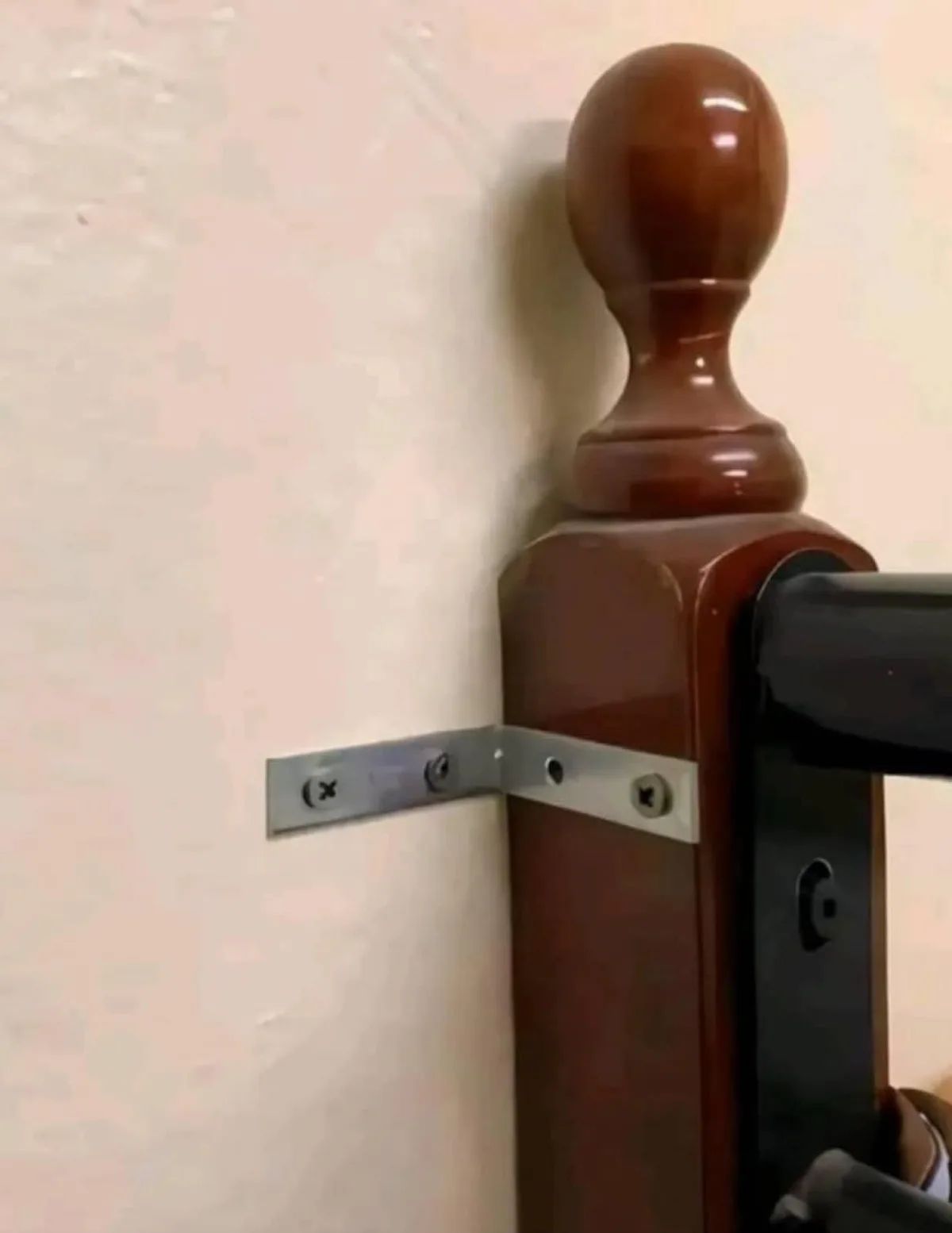 A close-up photo shows a shiny brown wooden stair railing post mounted to a light-colored wall with a metal L-bracket. The…