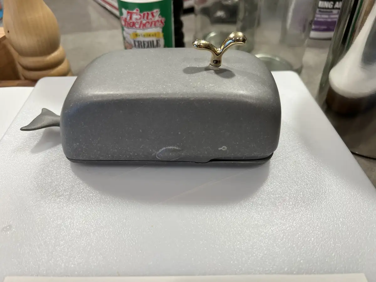 A gray, whale-shaped butter dish with a gold-colored handle sits on a white cutting board. The whale has a tail fin on its…