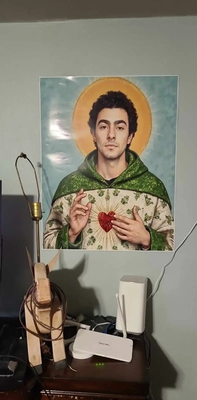 A poster of a man with a halo and a sacred heart, styled like a saint, is mounted on a wall above a desk with a router and a…