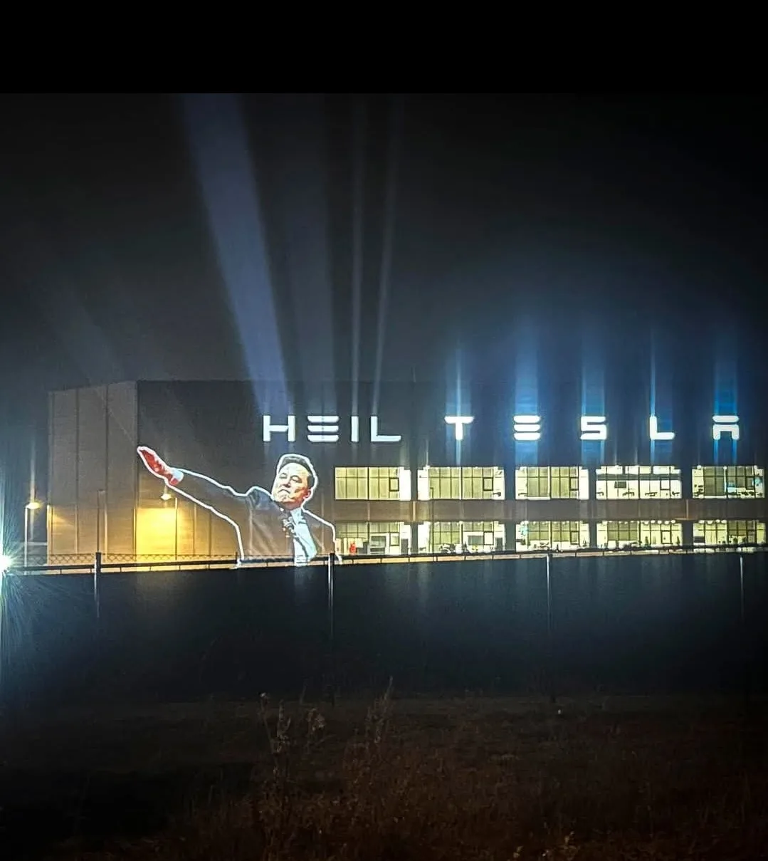 A nighttime photo of a large building with illuminated letters spelling 'HEIL TESLA' above its windows. Projected onto the…
