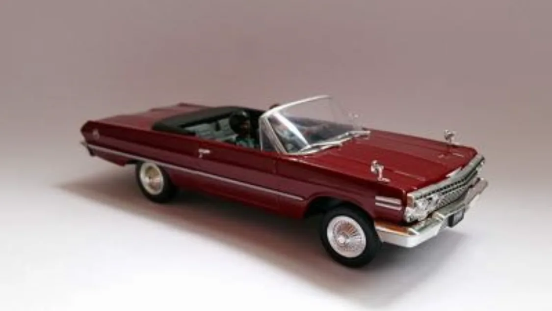 A maroon die-cast model of a classic American convertible car, shown from a three-quarter front angle against a plain white…