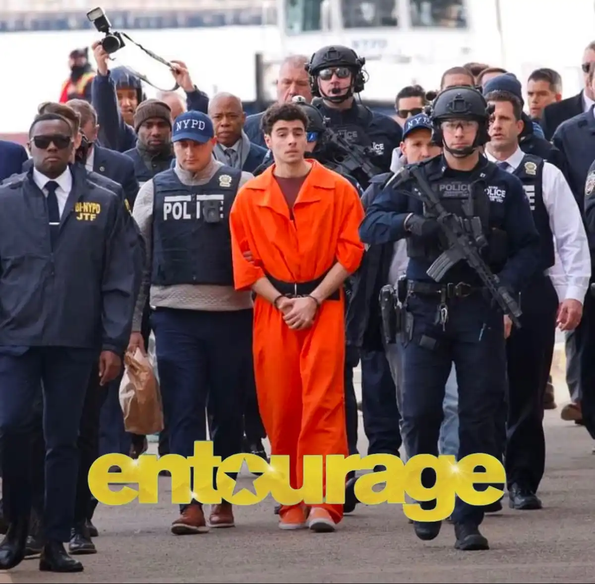 A man in an orange prison jumpsuit is being escorted by numerous NYPD officers, some in tactical gear and others in standard…