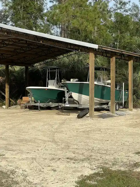Two green and white boats are parked side-by-side on trailers under a simple wooden and metal roof structure, surrounded by…