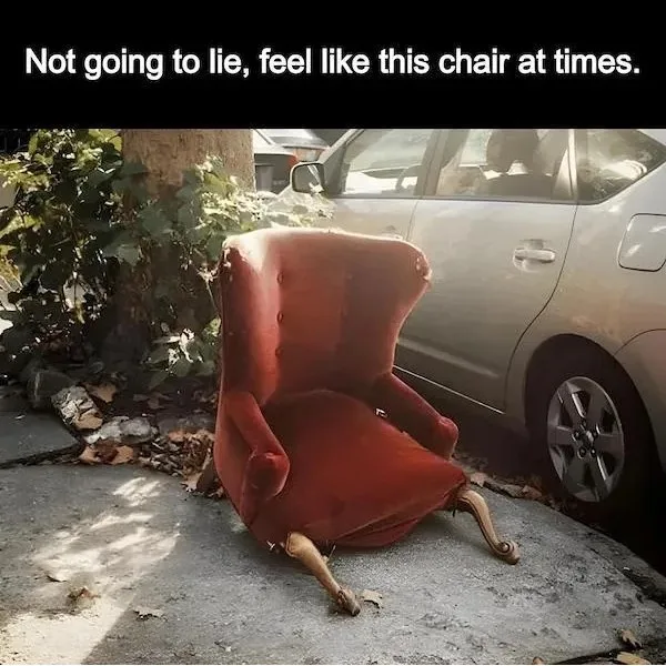 A red, upholstered wingback chair with ornate wooden legs is tipped over and abandoned on a concrete sidewalk next to a tree…