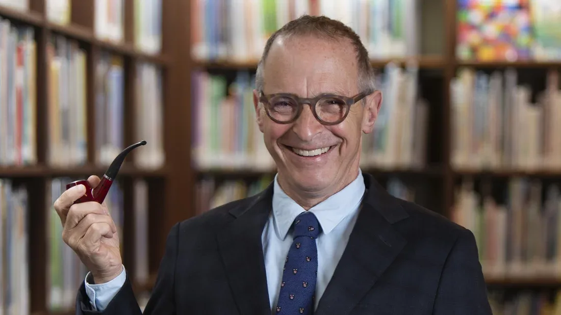 a smiling man in a suit and glasses holds a red pipe in front of bookshelves. The overlaid text reads 'i'm not a fan of the…