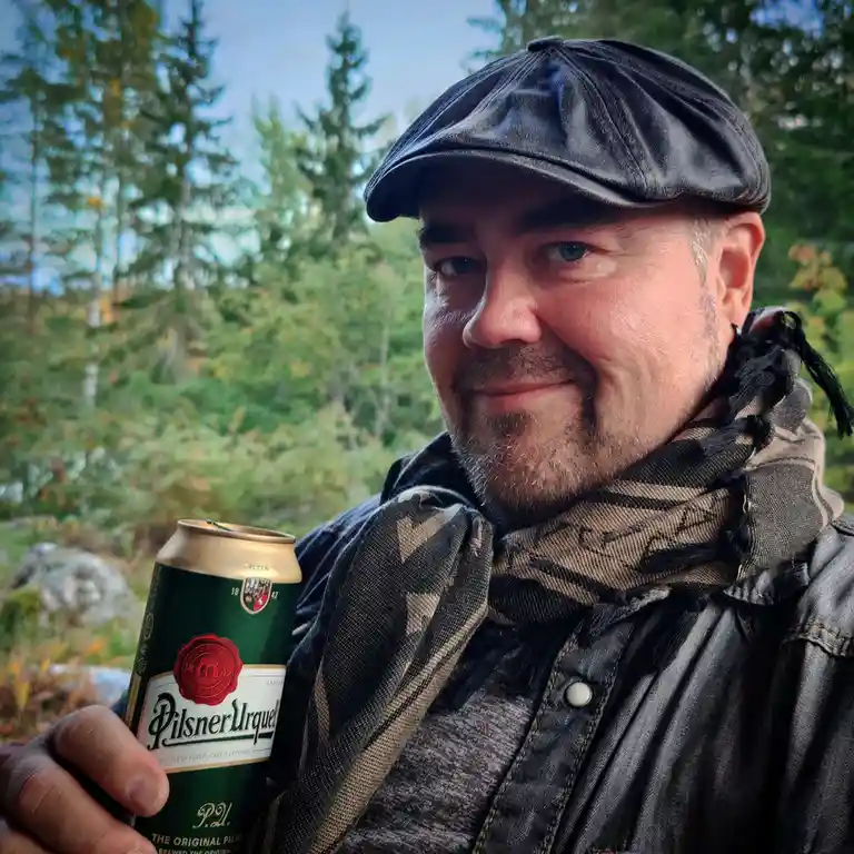 A man with a beard and a black flat cap holds a can of Pilsner Urquell beer outdoors in a forest setting. He wears a…