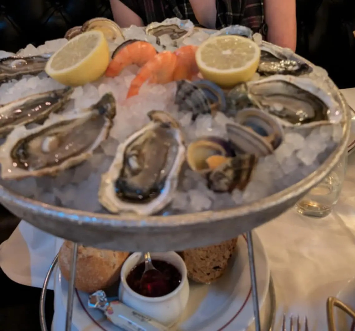 A two-tiered metal serving dish holds a seafood platter with raw oysters on the half shell, shrimp, and clams nestled in…