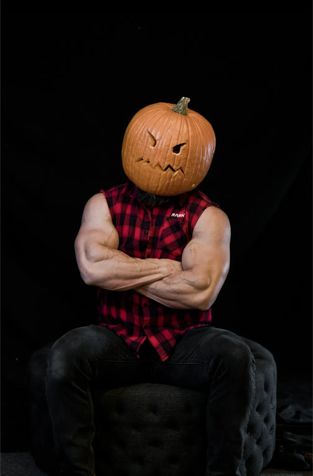 A muscular man with a beard sits on a dark tufted chair against a black background, arms crossed, wearing a red and black…