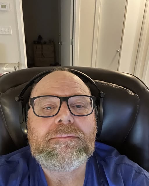 A middle-aged man with a beard and glasses sits in a black leather chair wearing headphones, looking directly at the camera…