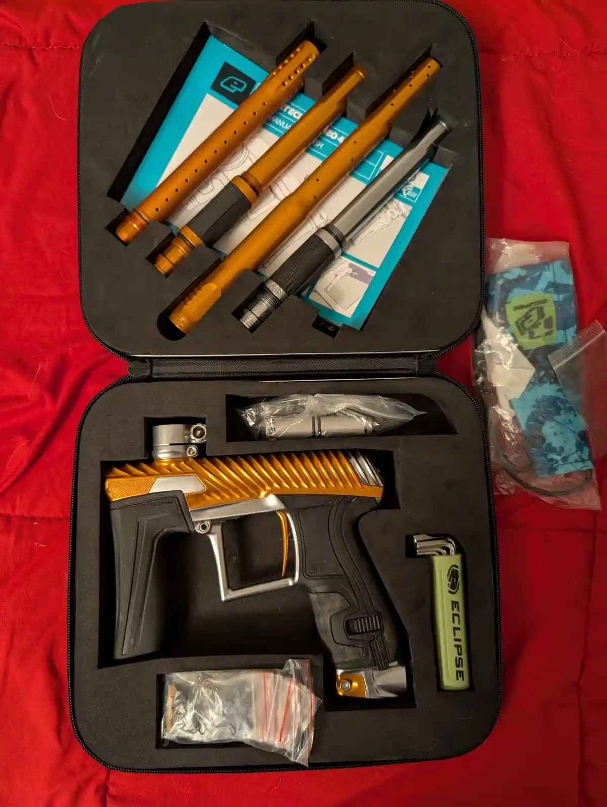 An open black foam-lined case on a red fabric surface contains a paintball marker with gold and black accents, several…