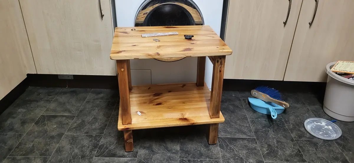 A wooden two-tier table stands in a laundry room, positioned directly in front of a front-loading washing machine. The top…
