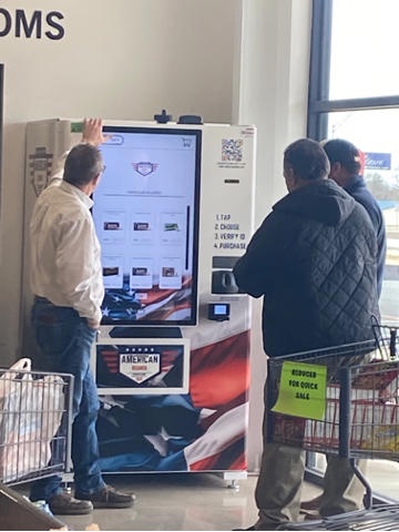 Three men stand before a large touchscreen kiosk in a store, one touching the screen while others wait. The kiosk displays a…