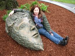 A woman is lying on the ground next to a large bronze statue head, smiling at the camera. She has one arm draped over the…