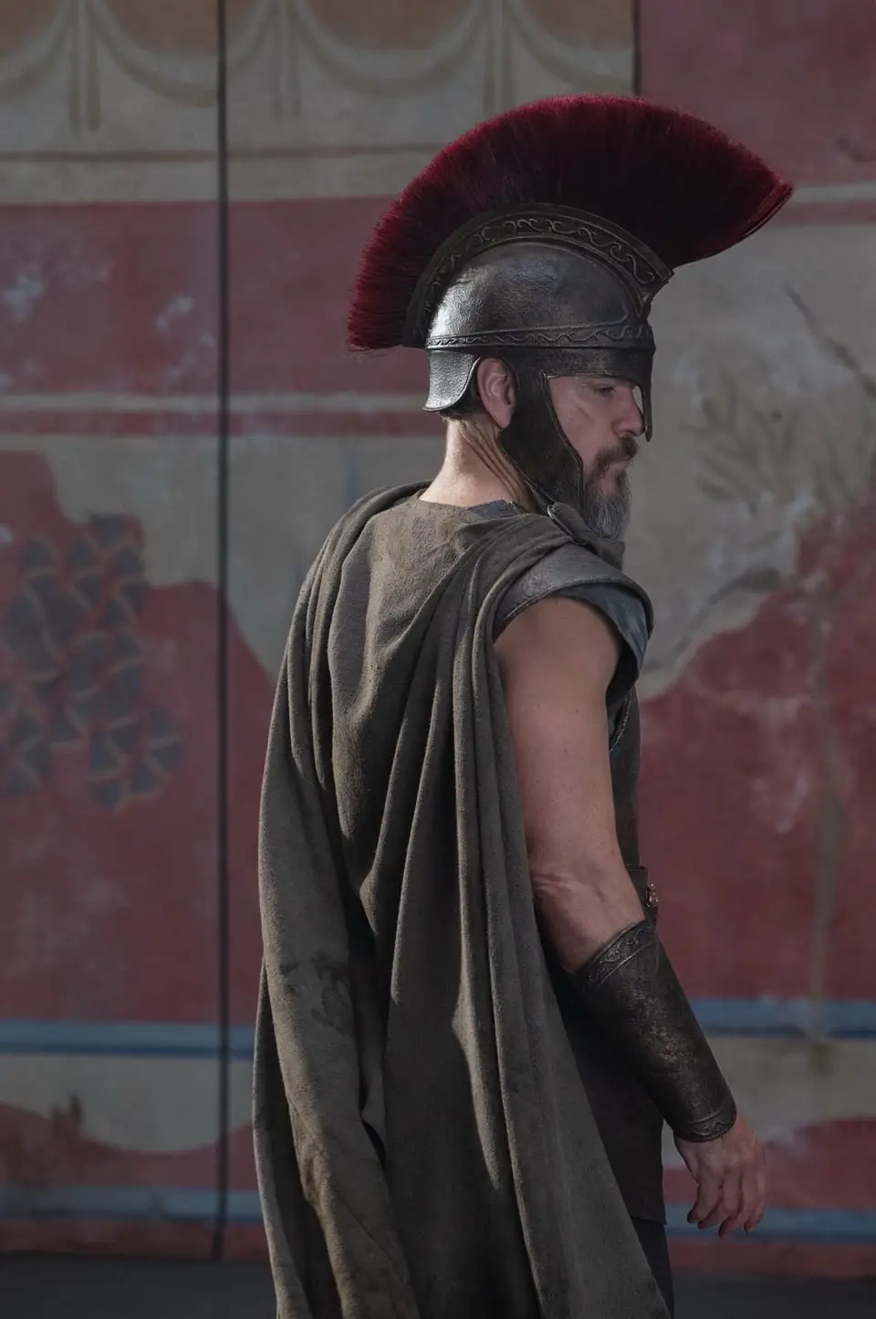 A man in ancient Roman-style armor, including a helmet with a red plume and a draped cloak, stands in profile looking away…