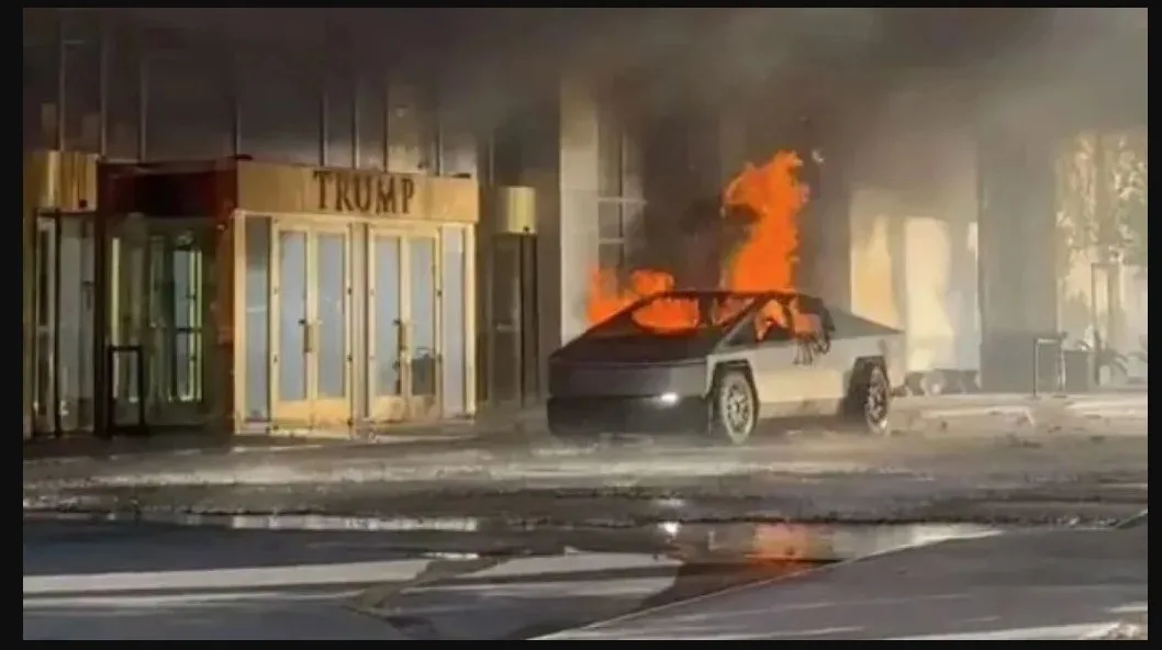A Tesla Cybertruck is engulfed in flames on a street in front of a building with a prominent 'TRUMP' sign above its…