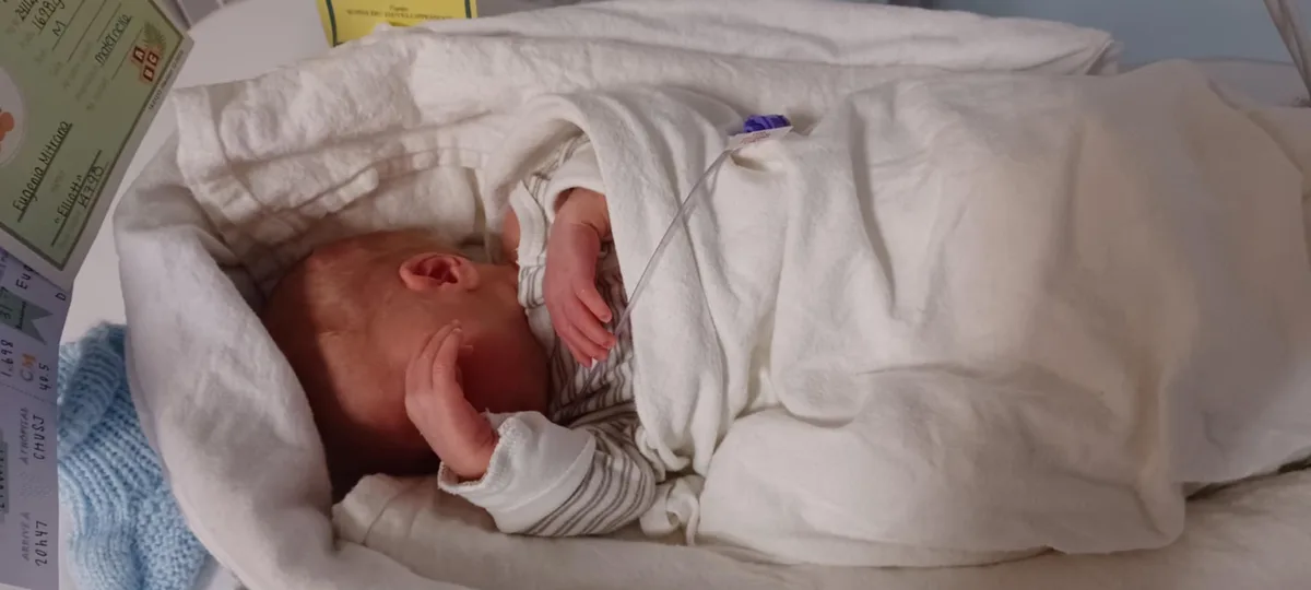 A newborn baby wrapped in a white blanket lies in a hospital bassinet, with a medical IV line taped to its arm. A hospital…