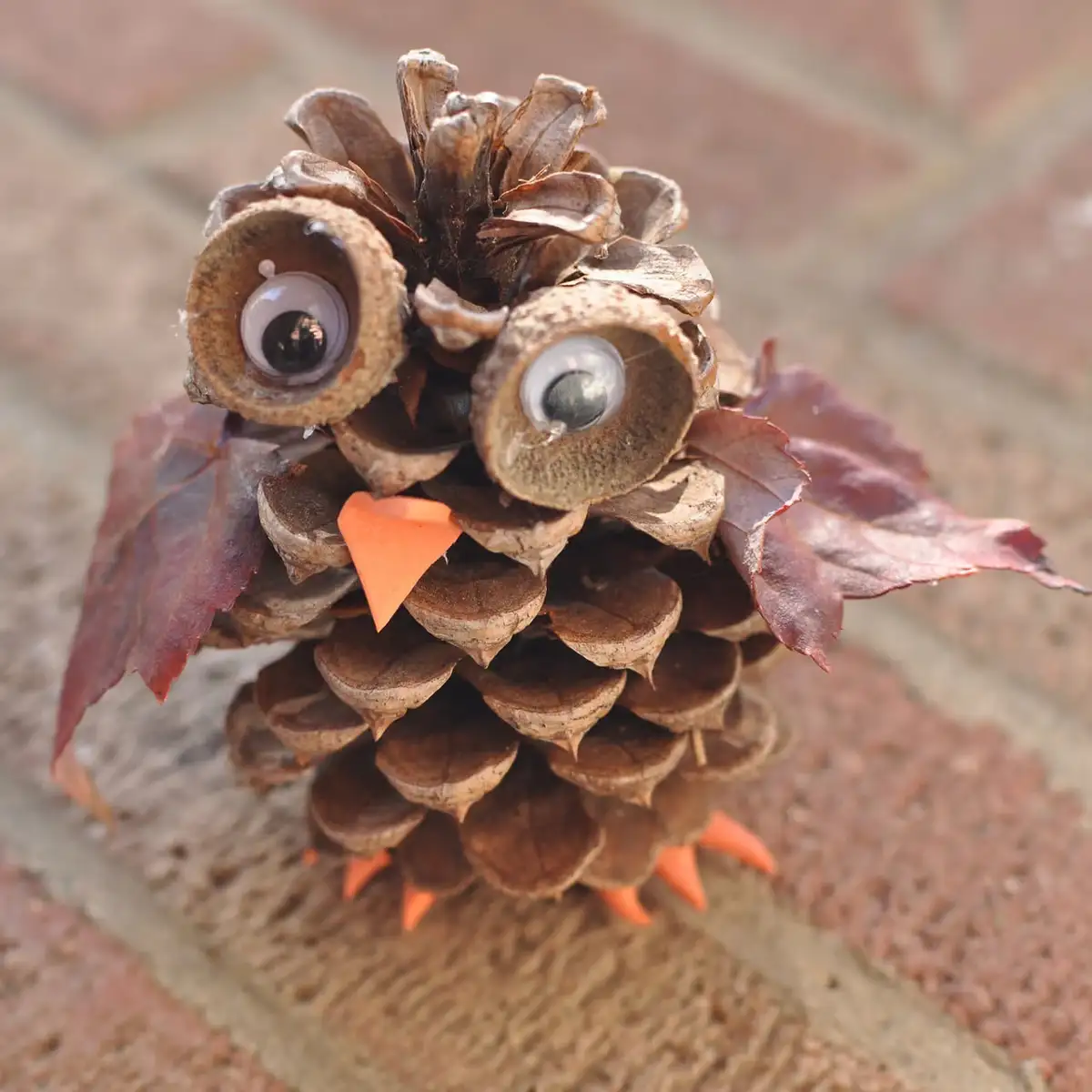 A pinecone has been creatively decorated with large googly eyes, an orange paper beak, and orange paper feet, with red…