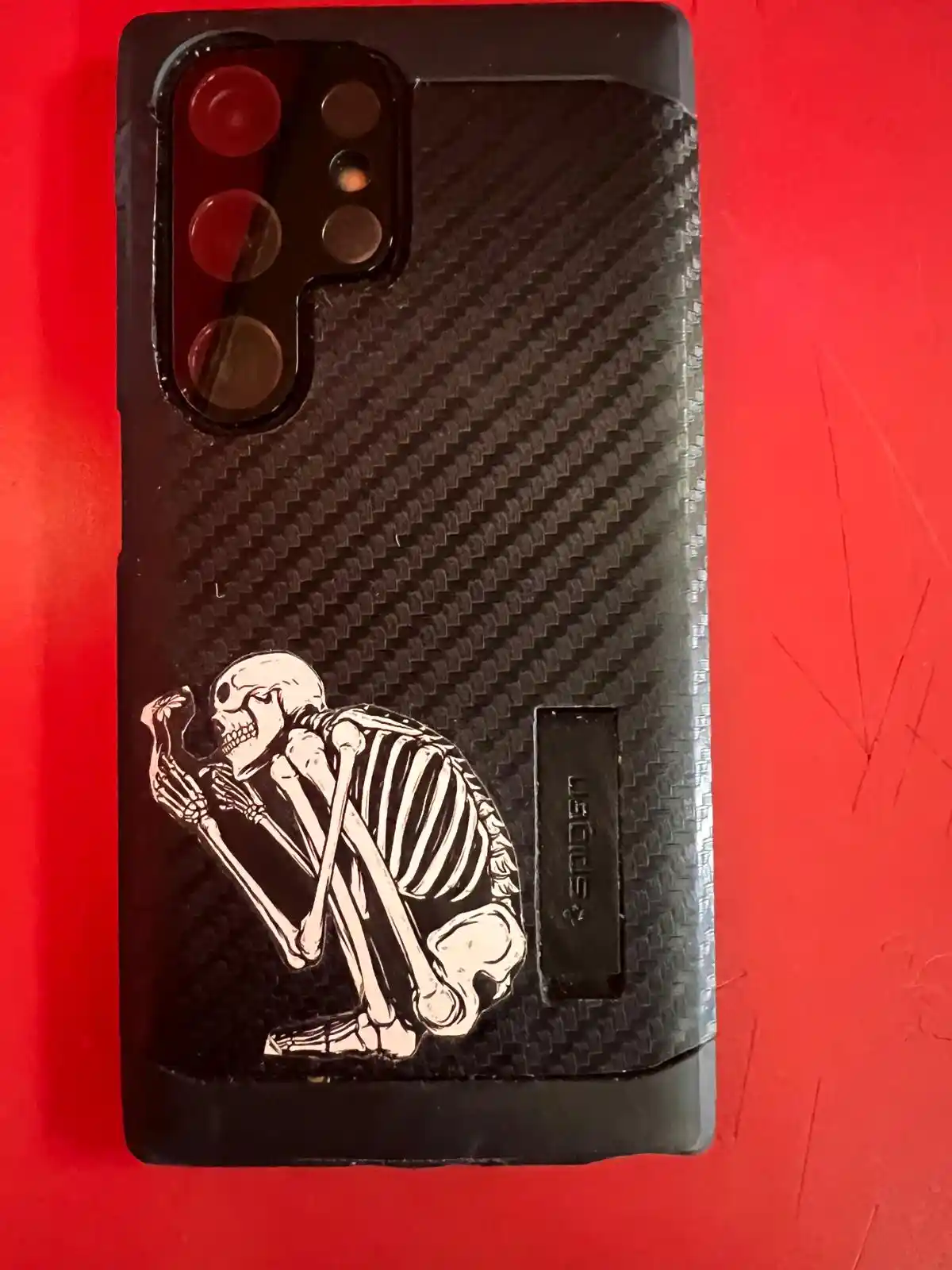 A black phone case with a carbon fiber texture and a white skeleton illustration of the Thinker pose is shown against a red…