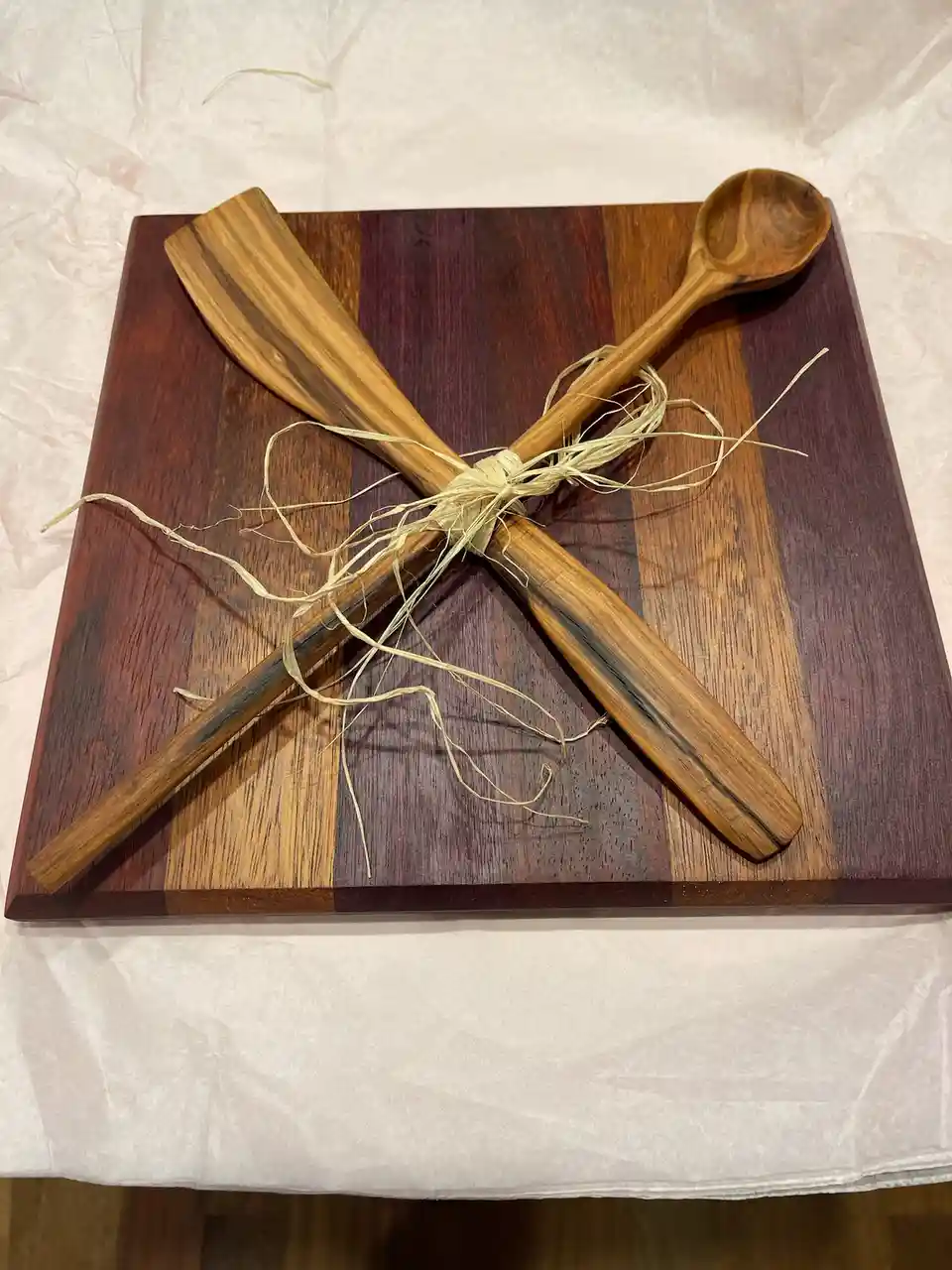 A wooden spoon and spatula are tied together with twine and placed on a striped wooden cutting board, which rests on a white…
