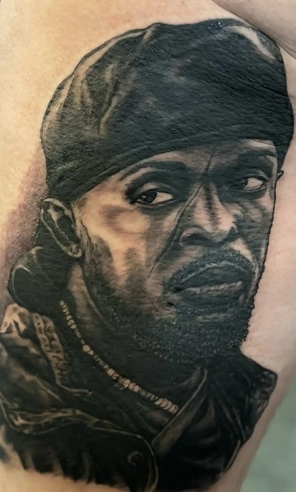 A detailed black and grey tattoo on human skin depicts a man with a headwrap, beard, and chain necklace, looking intensely…