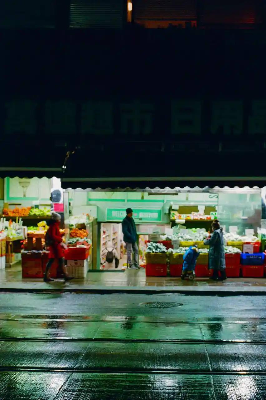 A nighttime photo of a wet street with a brightly lit grocery store, where three people are shopping. The store has green…