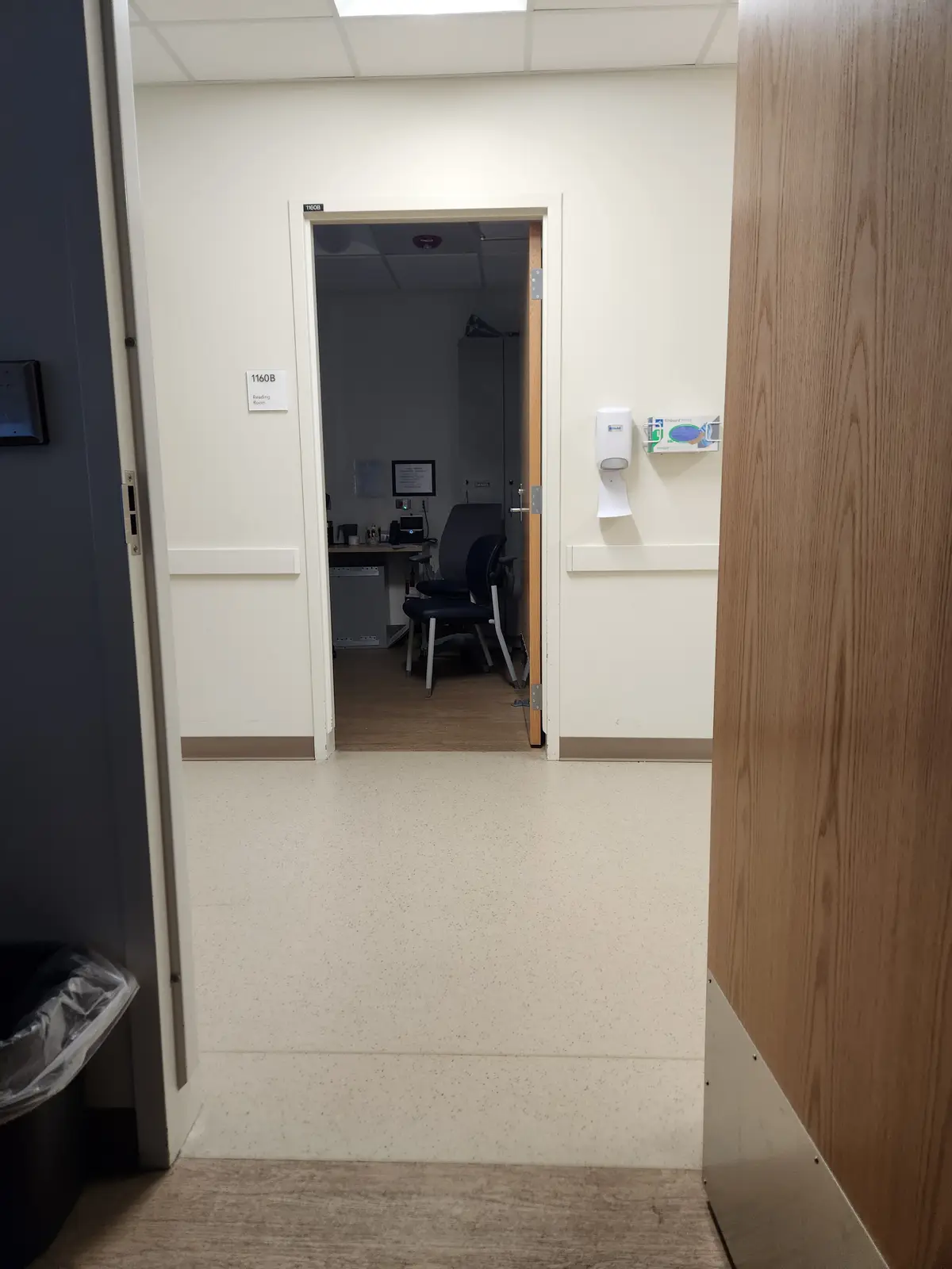 A view down a hallway into a room labeled '1160B Reading Room', which contains a desk, chairs, and medical equipment. The…