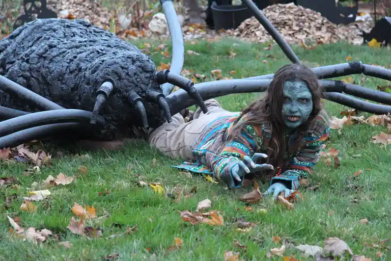 A person with blue face paint and fangs lies on grass next to a large, dark, spider-like sculpture with long legs. The…