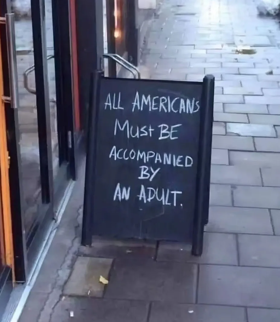 A black chalkboard sign stands on a wet sidewalk outside a glass door, written in white chalk. The sign reads 'ALL AMERICANS…