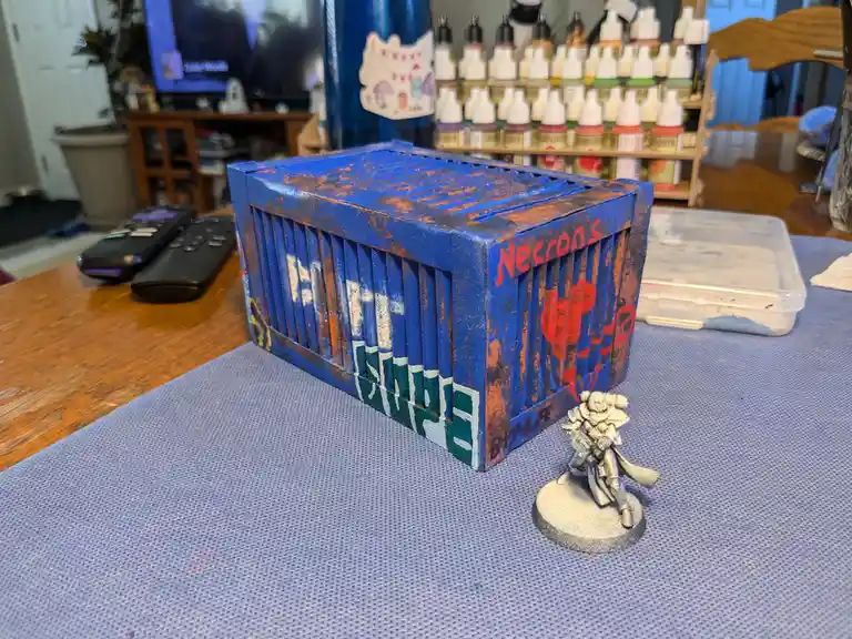 A hand-painted miniature blue shipping container with rust effects and red 'Necrons' text sits on a blue gaming mat, next to…