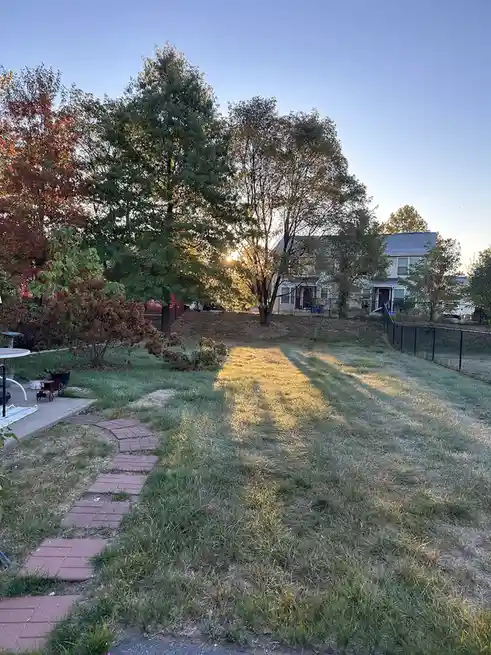 A backyard with a grassy lawn, a brick pathway, and trees showing autumn colors, with a house visible in the background. The…