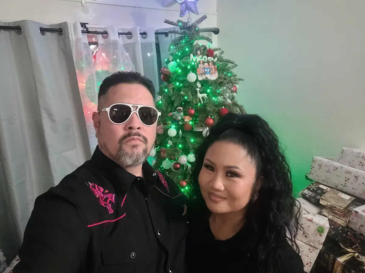 A man wearing white-framed sunglasses and a black shirt with pink embroidery stands beside a smiling woman in front of a…