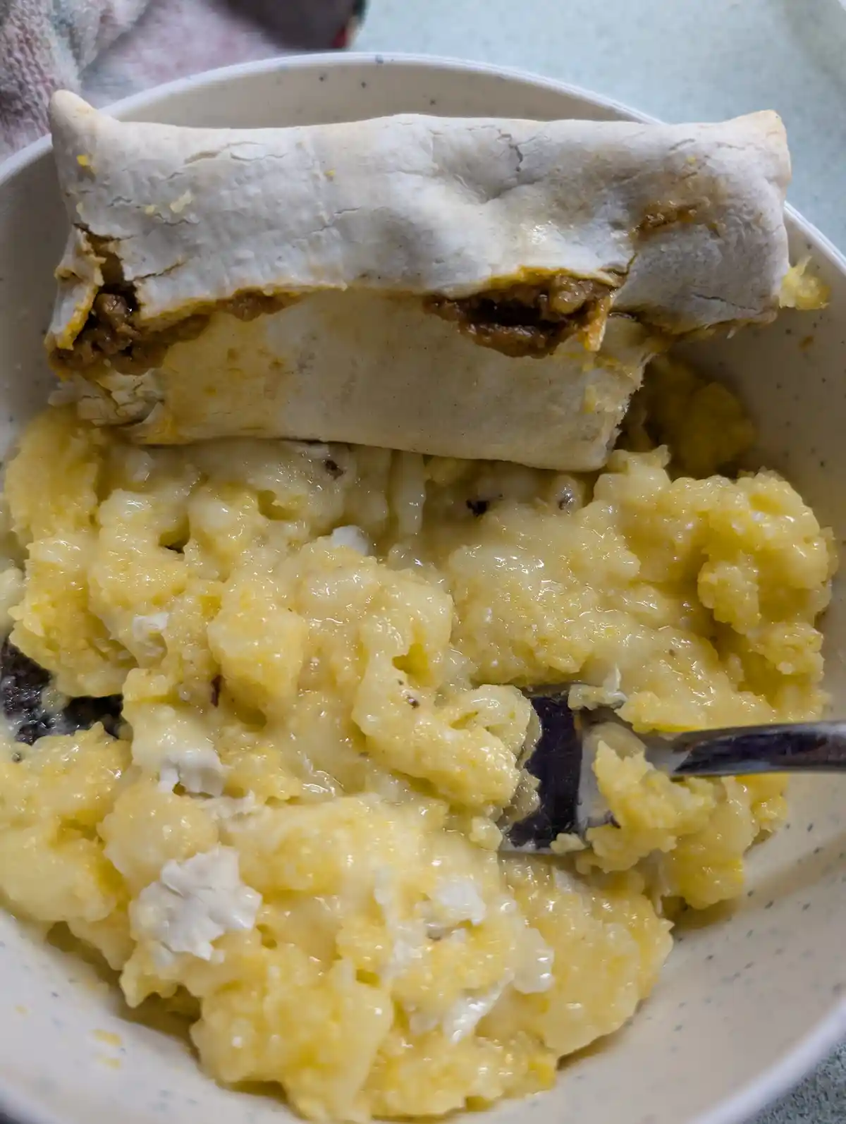 A close-up photo shows a partially eaten burrito resting on top of a bowl filled with scrambled eggs and melted cheese. A…