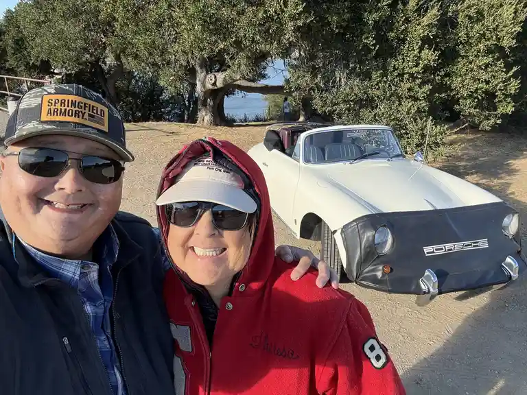 A smiling man and woman pose for a selfie outdoors next to a white vintage Porsche 356 convertible. The man wears a…