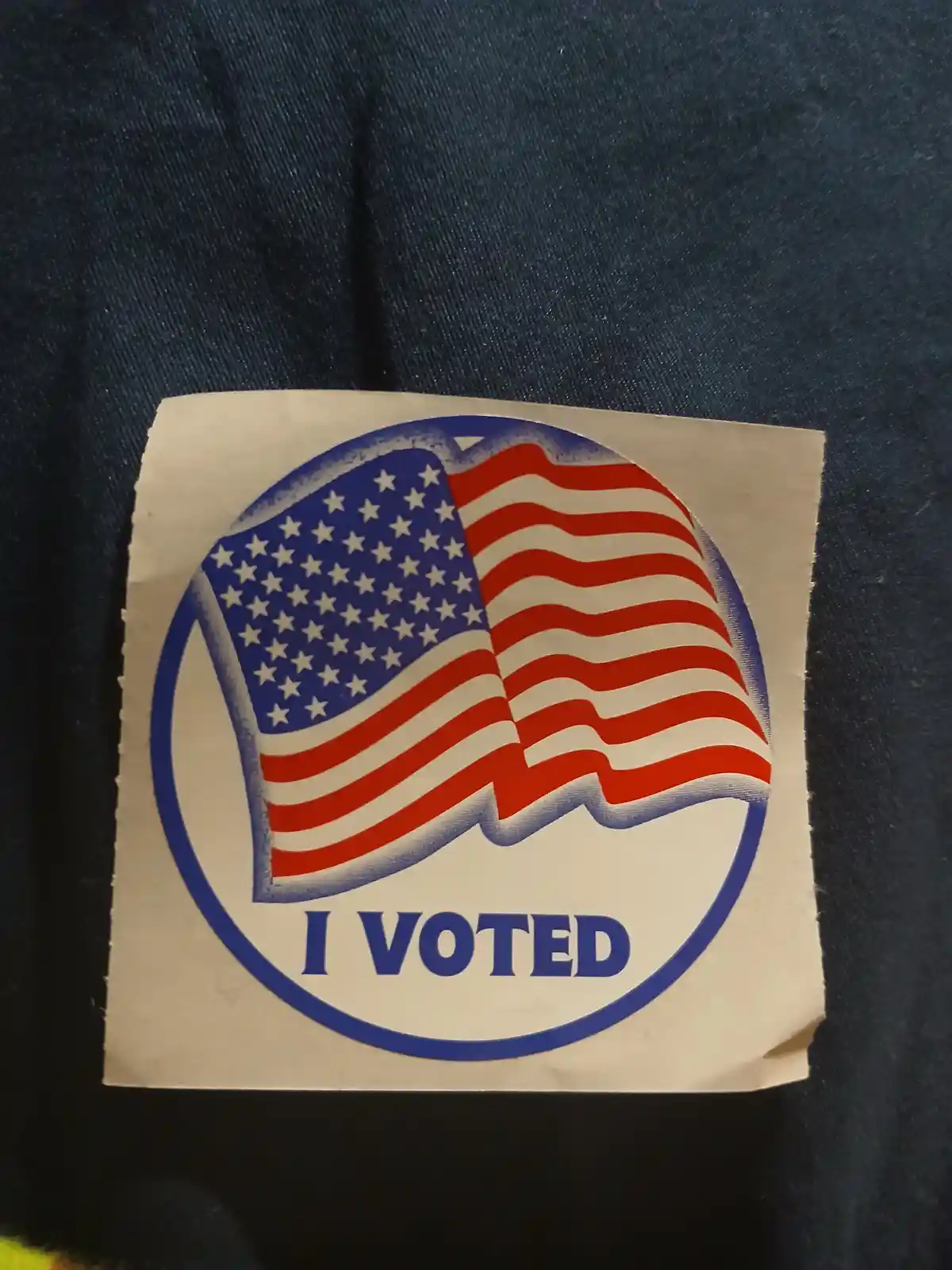 A close-up photo shows a worn, square-shaped 'I VOTED' sticker with a circular design. Inside the circle is a stylized…