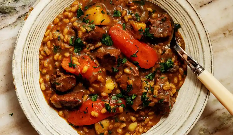 A close-up photo of a hearty bowl of beef stew with chunks of meat, carrots, potatoes, and barley in a rich brown gravy…