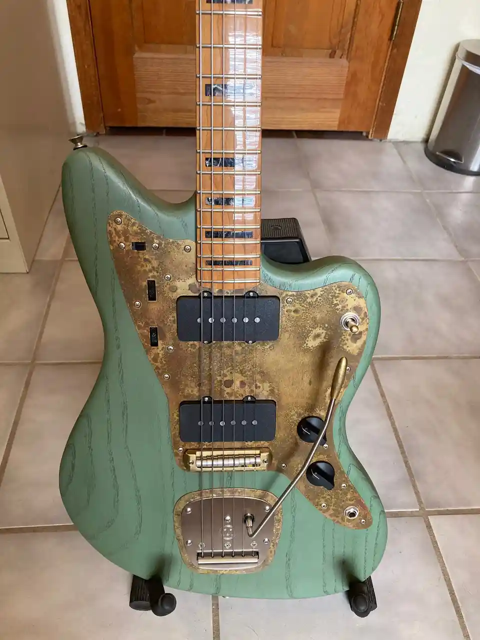A green electric guitar with a heavily rusted and weathered brass-colored pickguard and hardware stands upright on small…