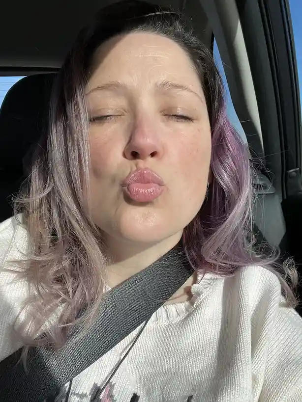 A person with purple and silver hair sits in a car seat, wearing a white sweater and a seatbelt, making a puckered lips face…