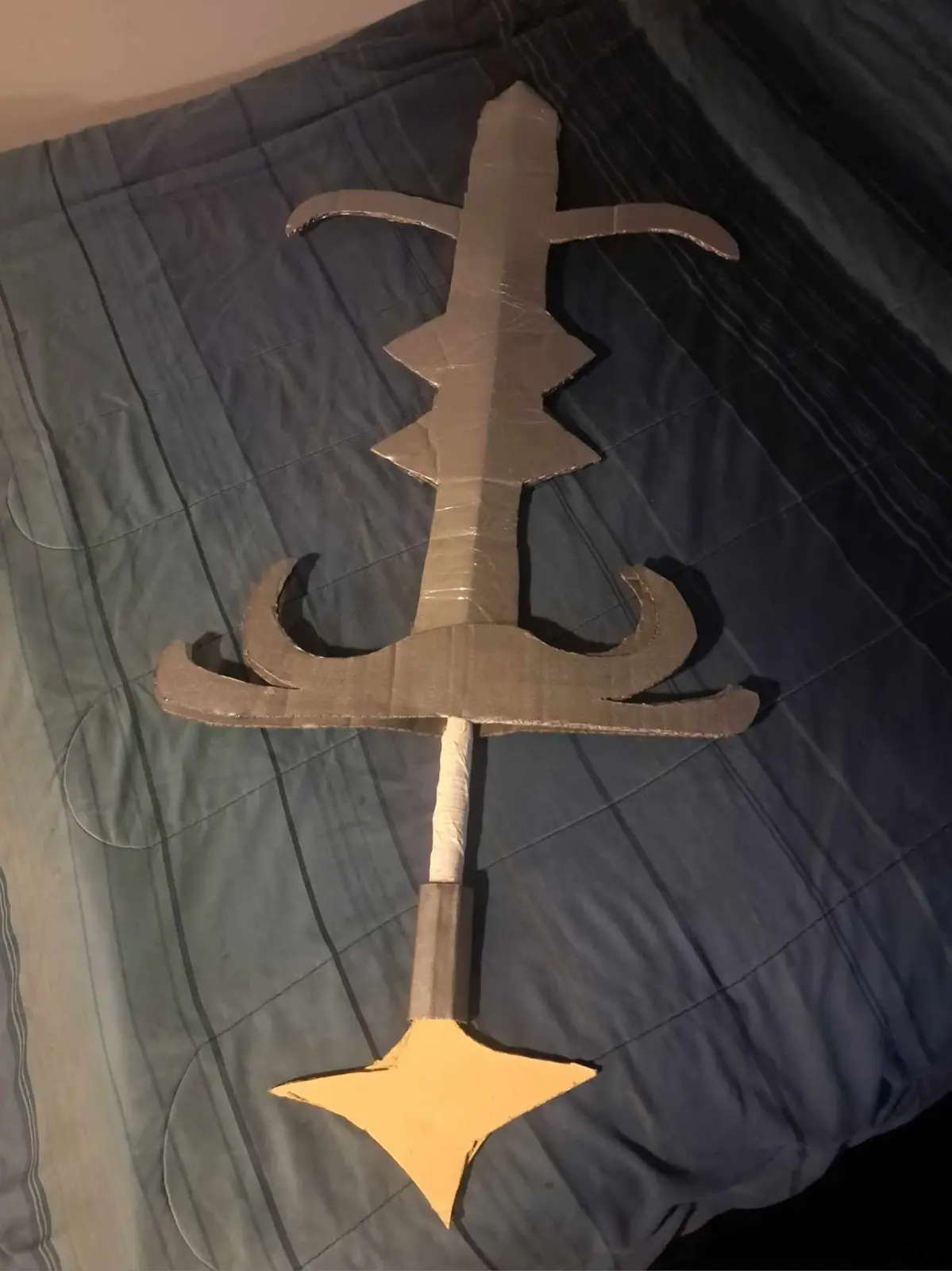 A handmade sword crafted from cardboard and duct tape lies on a dark blue quilted bedspread. The blade is long and jagged…