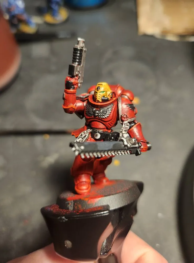 A hand holds a painted miniature of a red-armored warrior with a yellow helmet, holding a pistol in one hand and a…