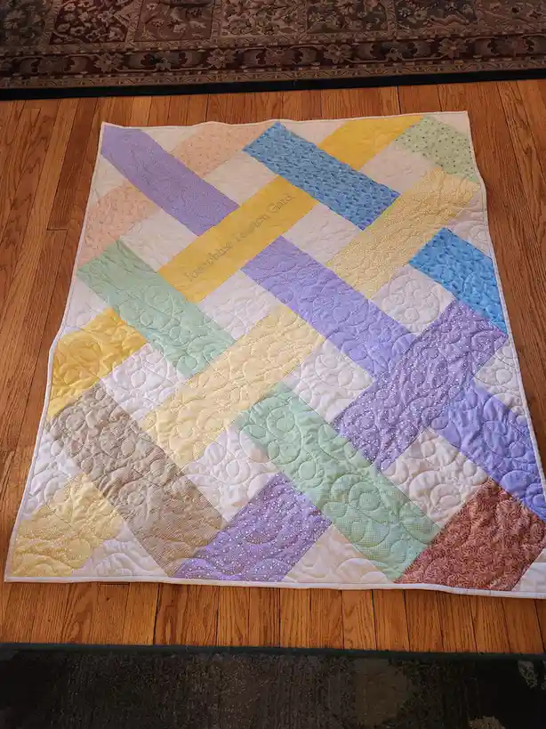 A handmade quilt with a diagonal patchwork pattern in pastel shades of yellow, blue, purple, and green lies on a wooden…