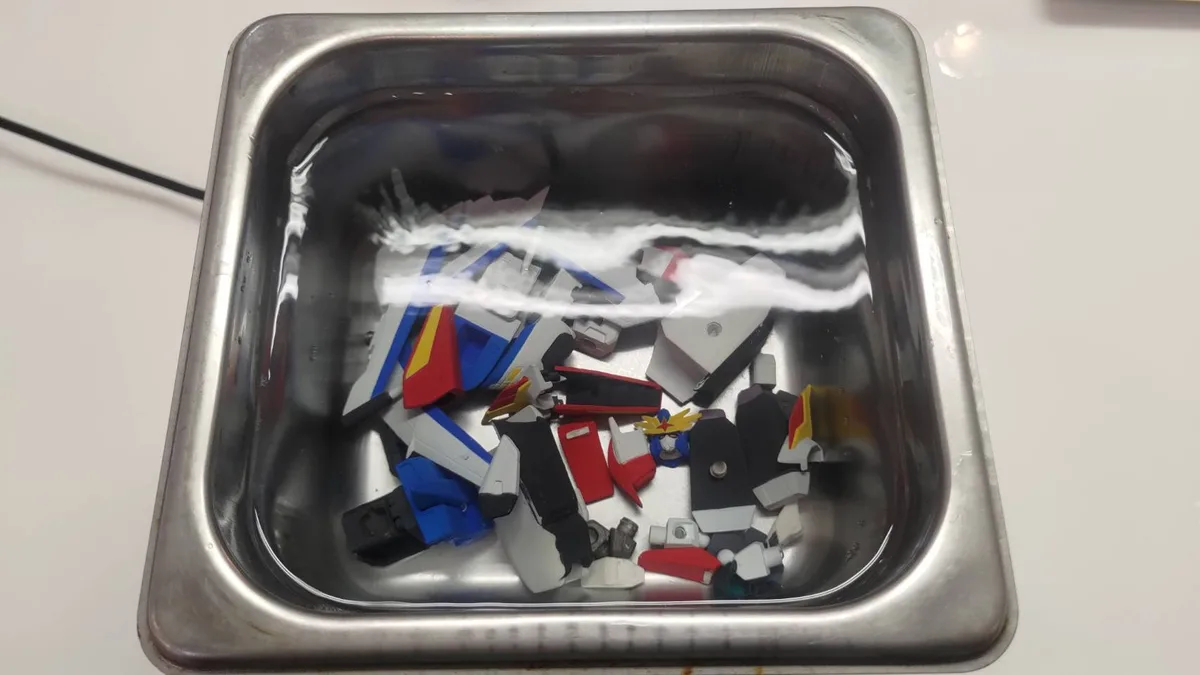 A stainless steel sink filled with water contains scattered, disassembled plastic parts of a Gundam model kit, including…