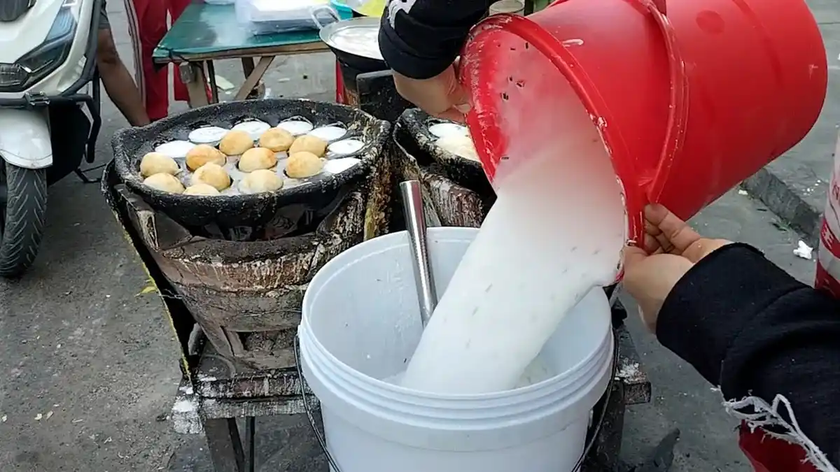 a person pours a thick white liquid from a red bucket into a white bucket, with a street food vendor's cooking station…